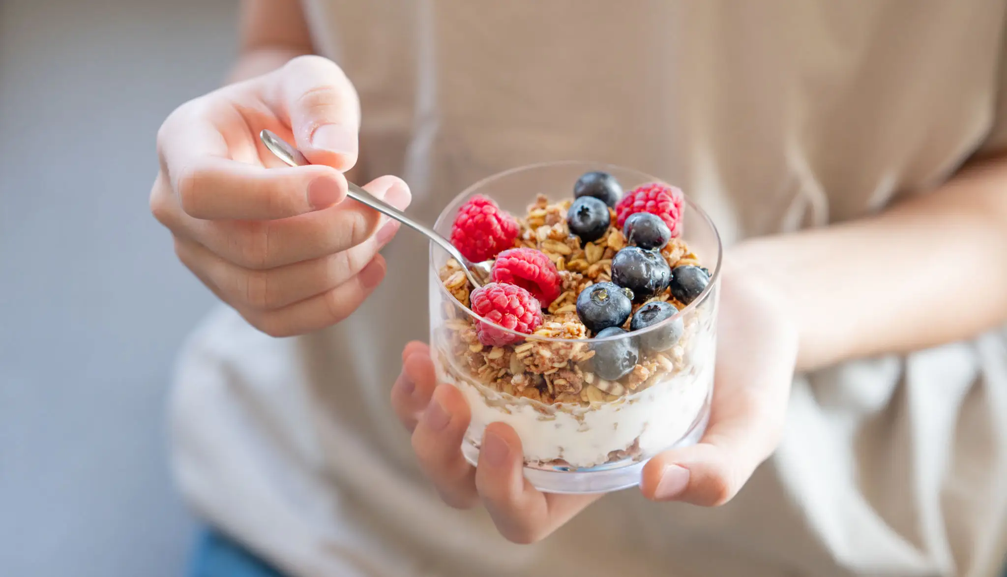 Hand holding a spoon and a glass of yogurt with granola and berries.