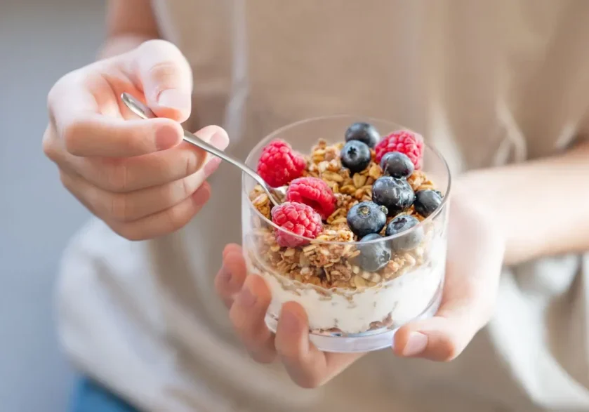 Hand holding a spoon and a glass of yogurt with granola and berries.